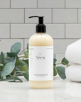 Sans Skincare Moisturizing Body Wash in cream-colored pump bottle displayed on a bathroom counter with eucalyptus leaves and folded white towels in the background