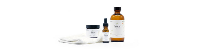 Sans Skincare product trio including a jar of Whipped Face Butter, a dropper bottle of Nourish Face Serum, and a large amber bottle of Nourishing Oil Cleanser, displayed with a white textured cloth