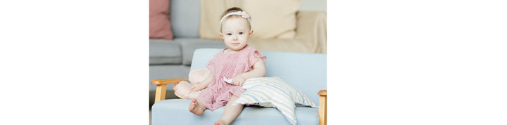 Baby sitting on a light blue chair, wearing a pink outfit with a floral headband, holding a small cloth, with pillows around
