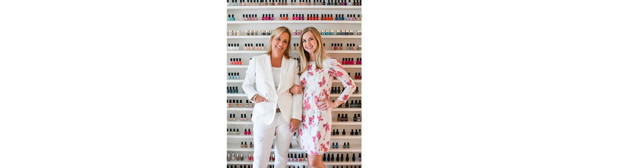 Two smiling women standing in front of a wall display of colorful nail polish bottles, one wearing a white suit and the other in a white floral dress