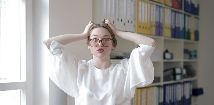 Young woman in red glasses and white blouse holding her head with both hands, making a surprised expression in an office setting